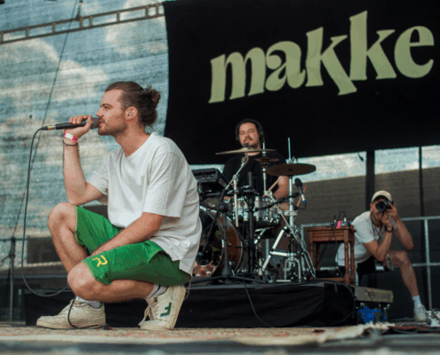 Ein Mann singt auf der Bühne. Er kniet und hält ein Mikrofon. Hinter ihm sind noch zwei Männer mit Musikinstrumenten.