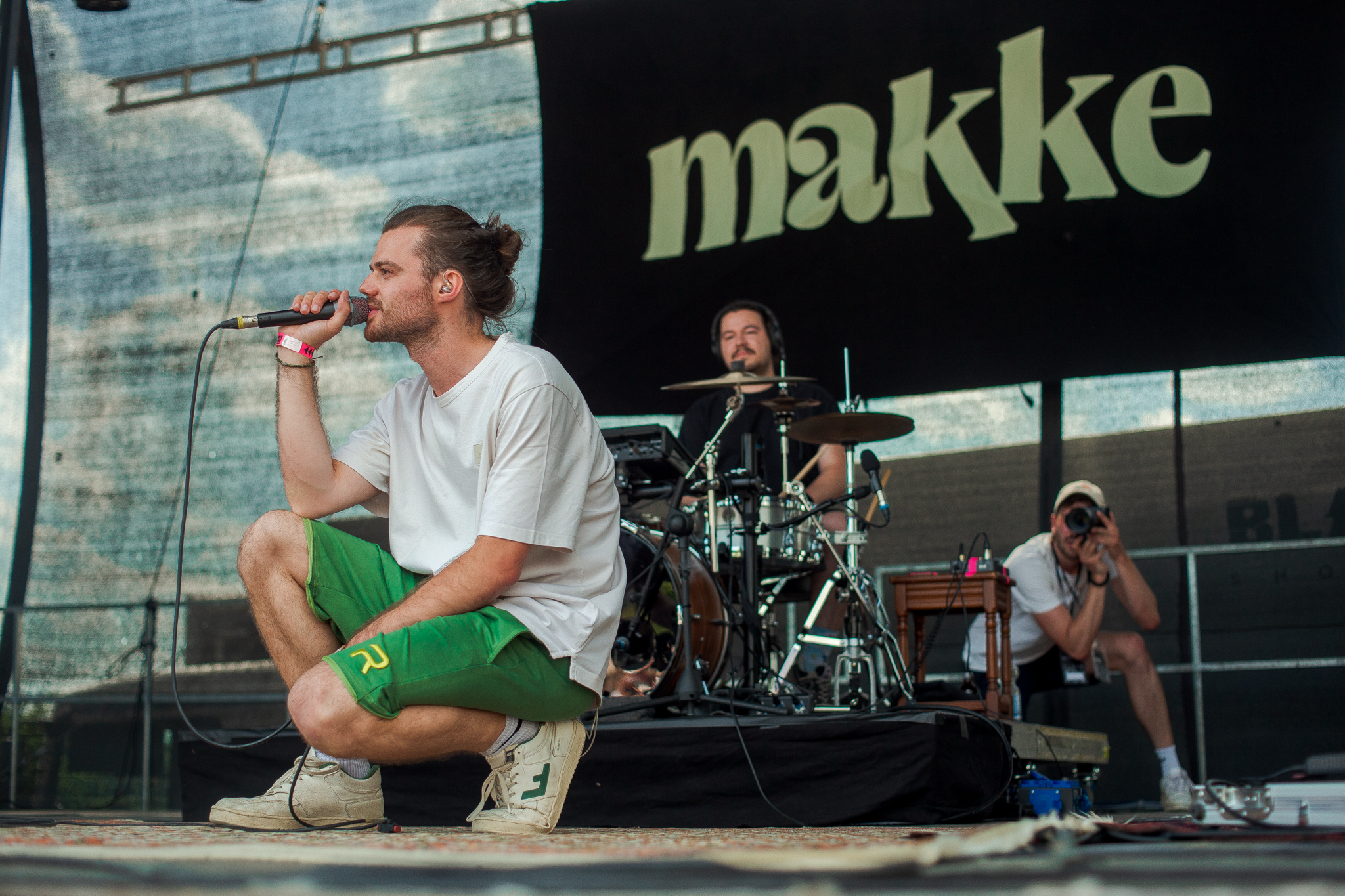 Ein Mann singt auf der Bühne. Er kniet und hält ein Mikrofon. Hinter ihm sind noch zwei Männer mit Musikinstrumenten.