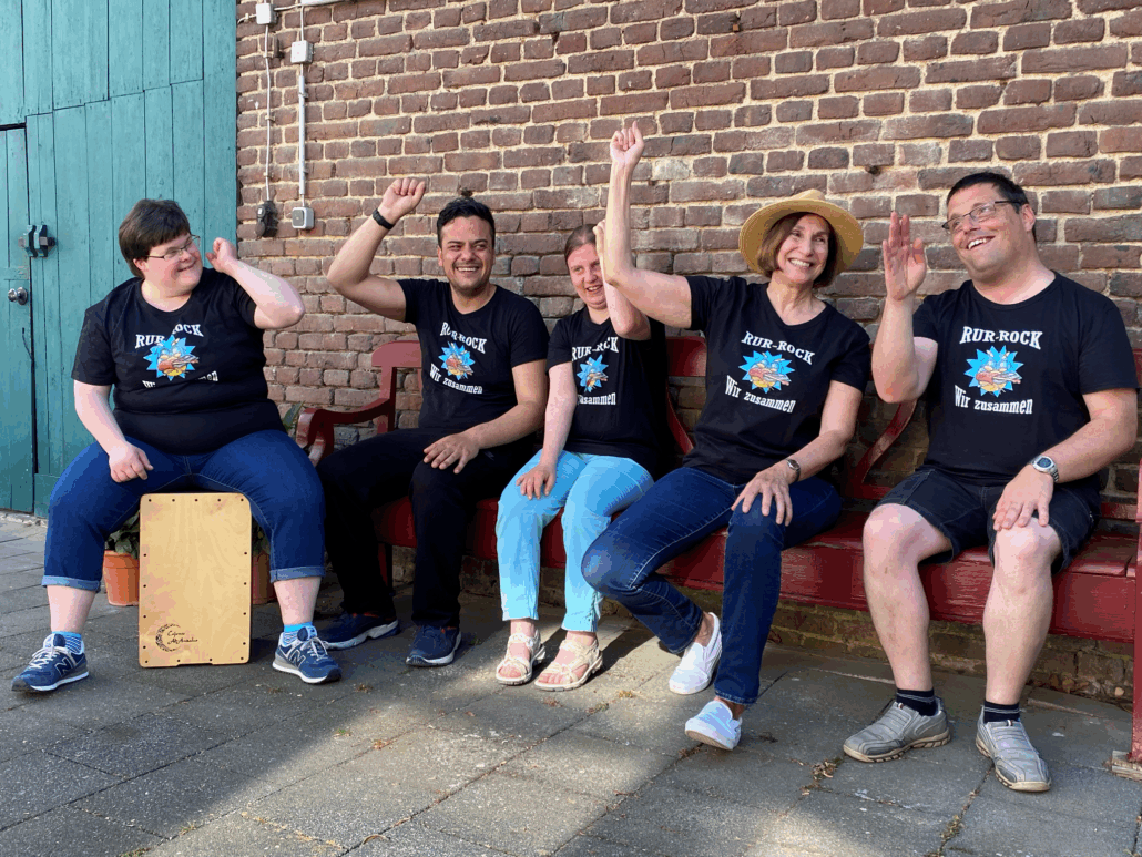 Zwei Personen sitzen vor einer Mauer. Die eine hält ein Holzbrett. Beide lächeln und zeigen Daumen hoch.