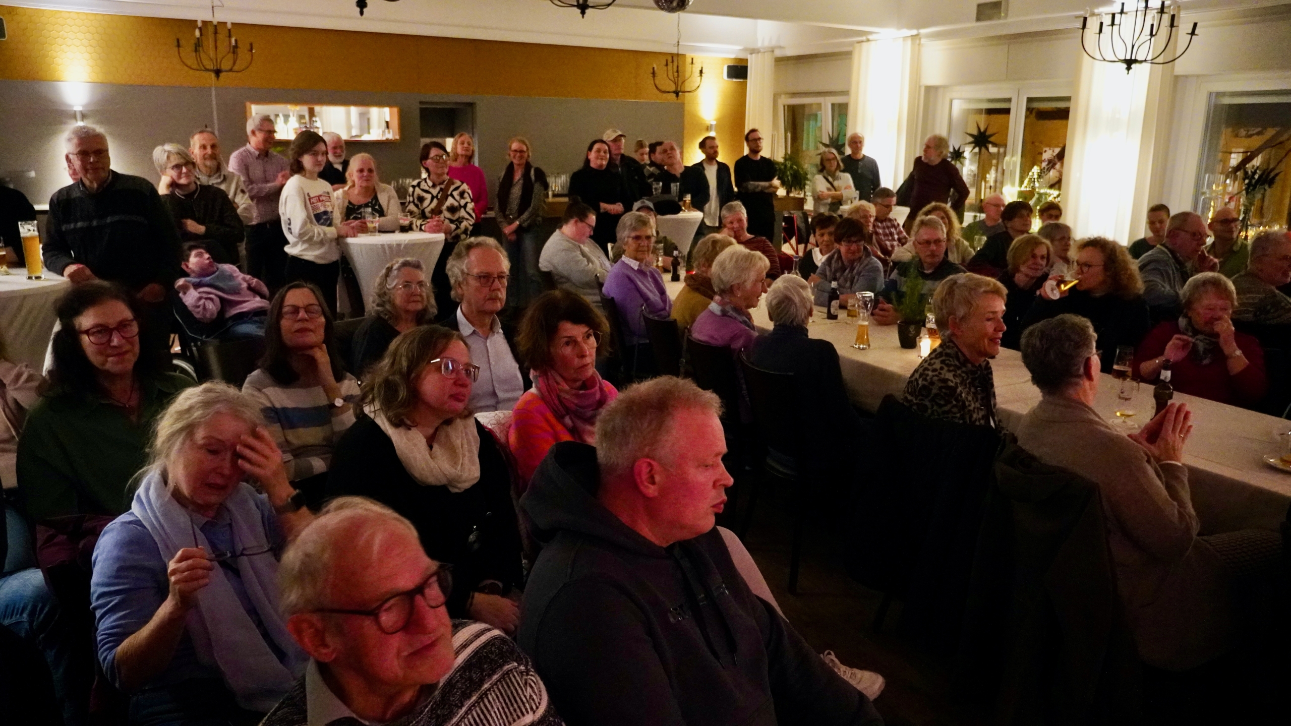 Viele Menschen sitzen an Tischen. Es sieht aus wie eine Feier. Es sind Lichter und Dekorationen zu sehen.