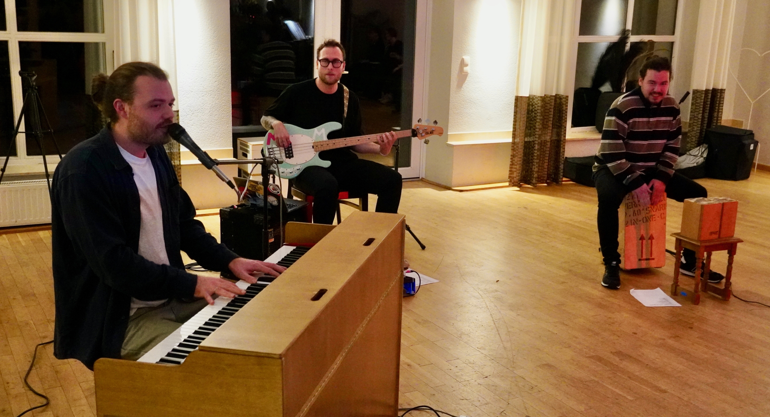 Drei Männer sitzen in einem Raum. Ein Mann spielt Klavier, die anderen sitzen auf Stühlen und halten Musikinstrumente.