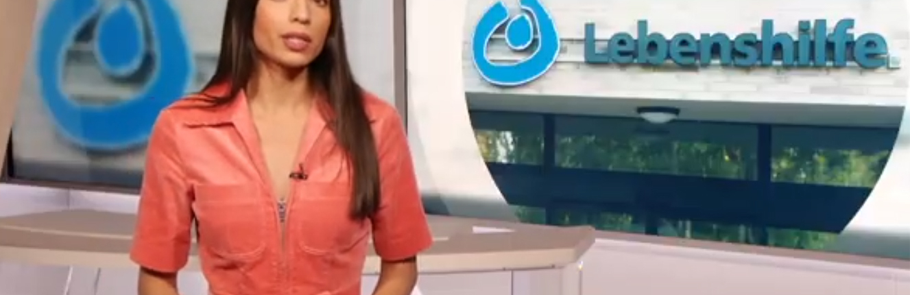 Eine Frau steht vor einem roten Hintergrund. Sie trägt ein rotes Oberteil und eine Uhr am Handgelenk.