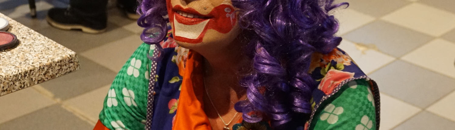 Ein Clown sitzt auf einem Stuhl. Er hat lila Haare und ein rotes Häubchen. Er lächelt und hat bunte Kleidung an.