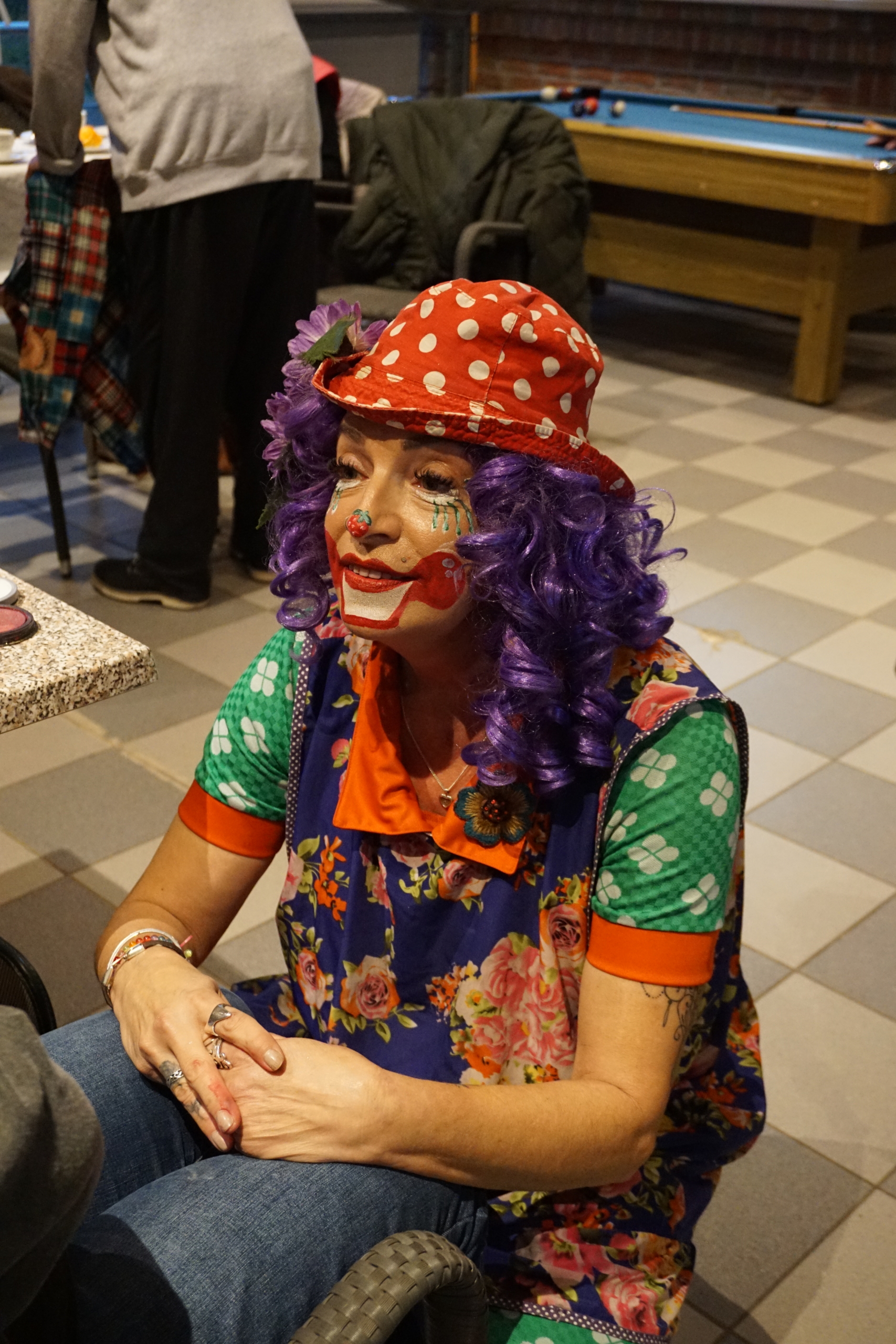 Ein Clown sitzt auf einem Stuhl. Er hat lila Haare und ein rotes Häubchen. Er lächelt und hat bunte Kleidung an.