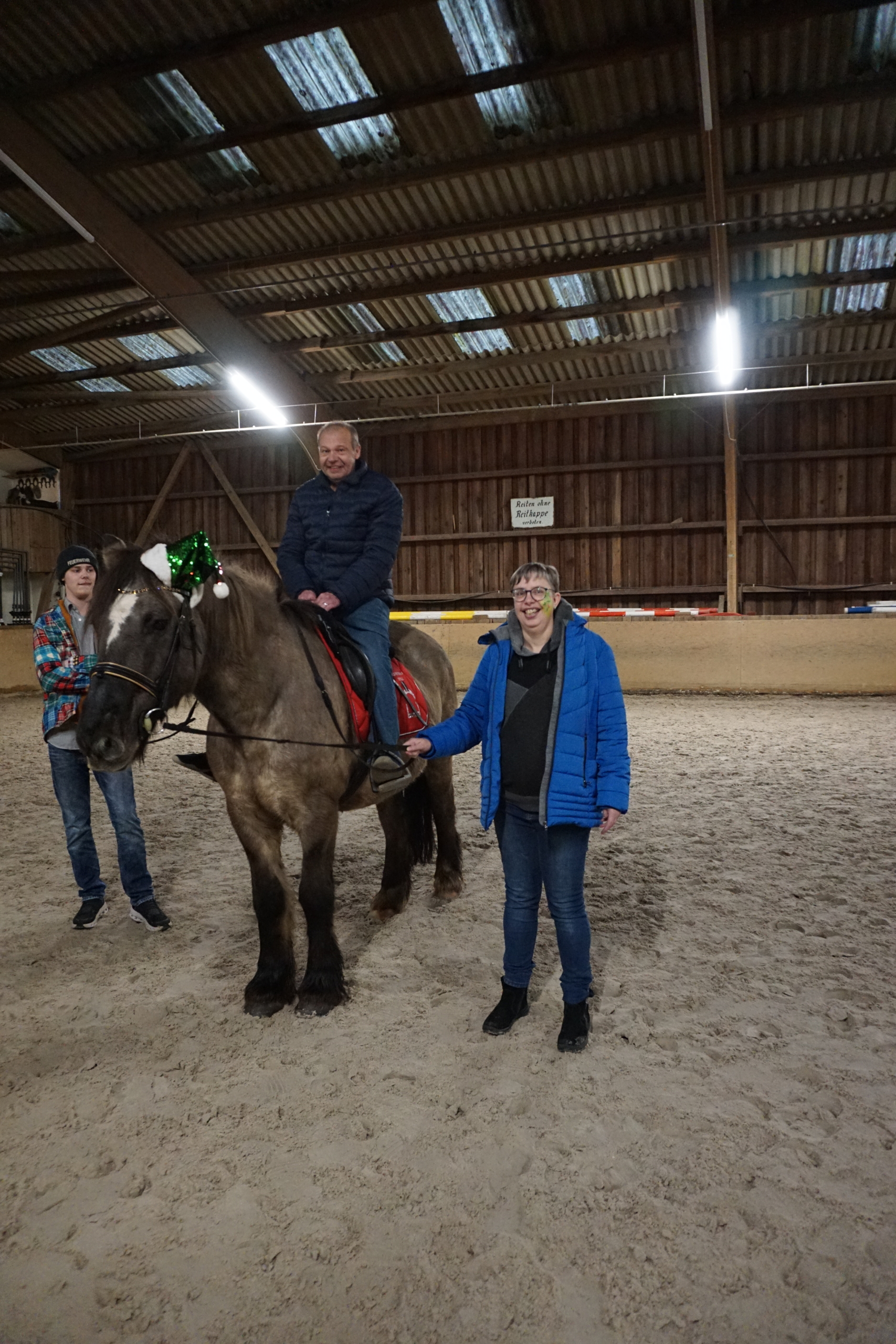 Ein Mann sitzt auf einem Pferd. Zwei Frauen stehen daneben. Sie sind in einem Stall.