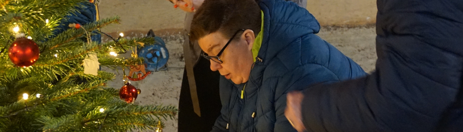 Zwei Personen hängen Weihnachtsdeko an einen Baum. Der Baum steht im Freien.