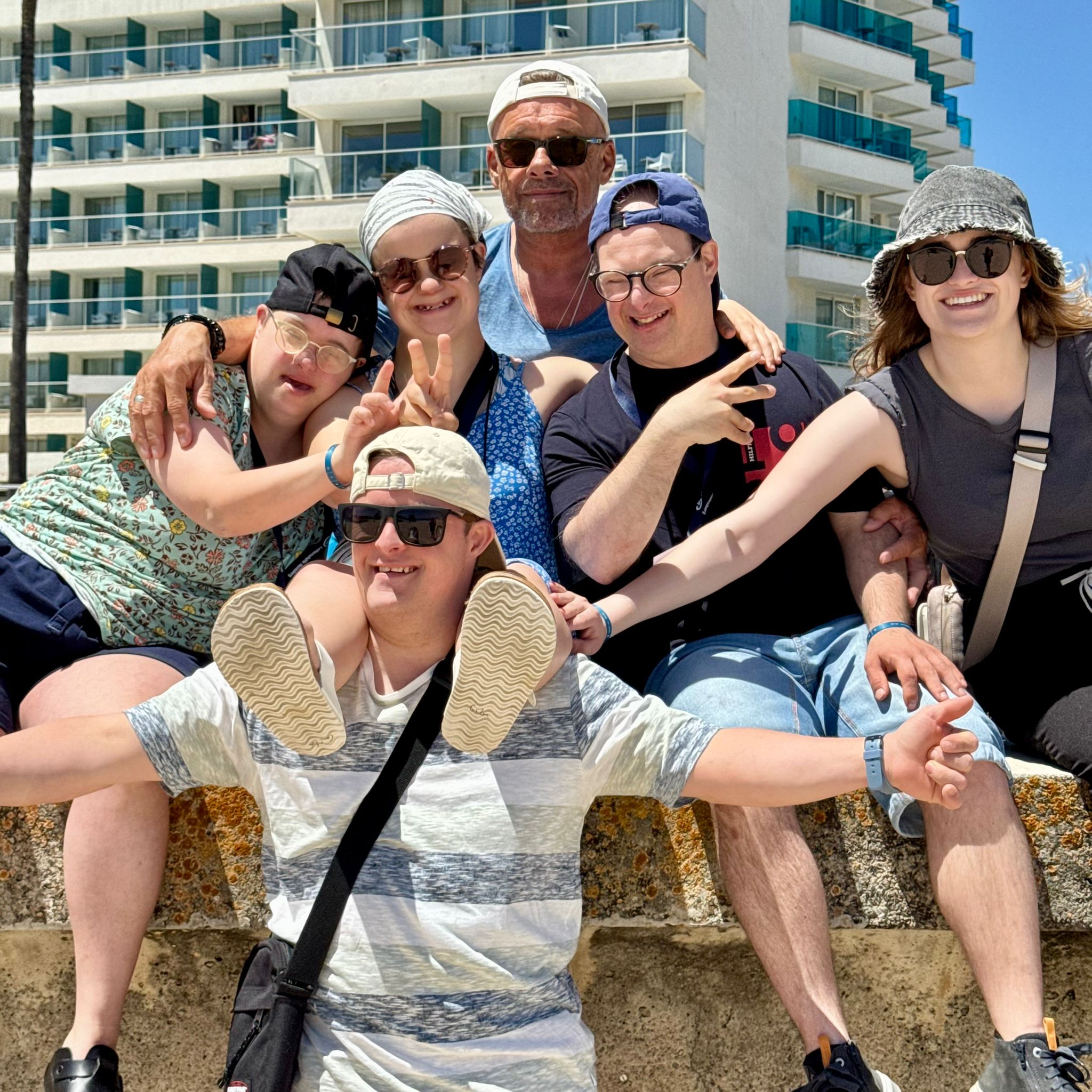 Zwei Männer sitzen auf einer Mauer am Meer. Einer trägt eine Kappe, der andere einen Hut. Hinter ihnen sind hohe Gebäude zu sehen.