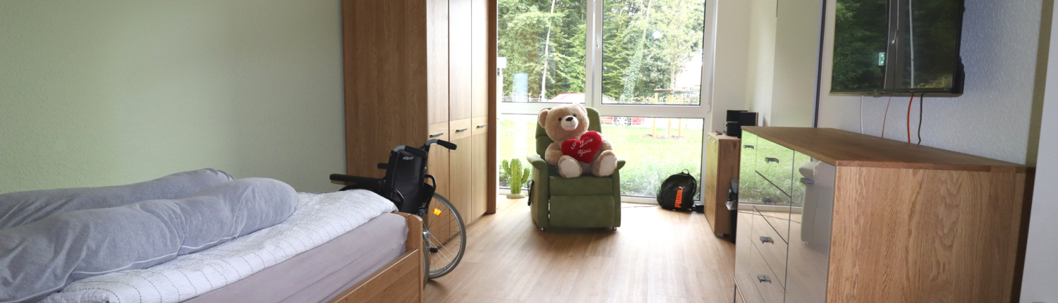 Ein helles Zimmer mit einem Bett und einem Schrank. Ein Teddybär sitzt auf einem Stuhl.