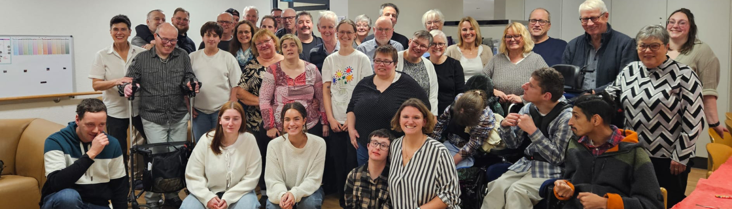 Viele Menschen stehen und sitzen in einem Raum. Sie lächeln und schauen in die Kamera.