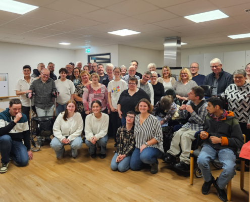 Viele Menschen stehen und sitzen in einem Raum. Sie lächeln und schauen in die Kamera.