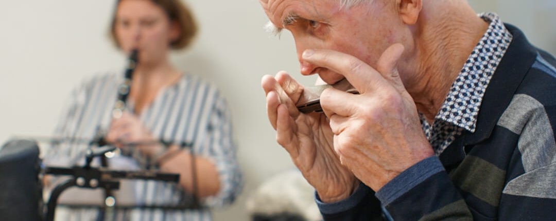 Ein Mann spielt Mundharmonika. Im Hintergrund steht eine Frau mit einem Instrument.