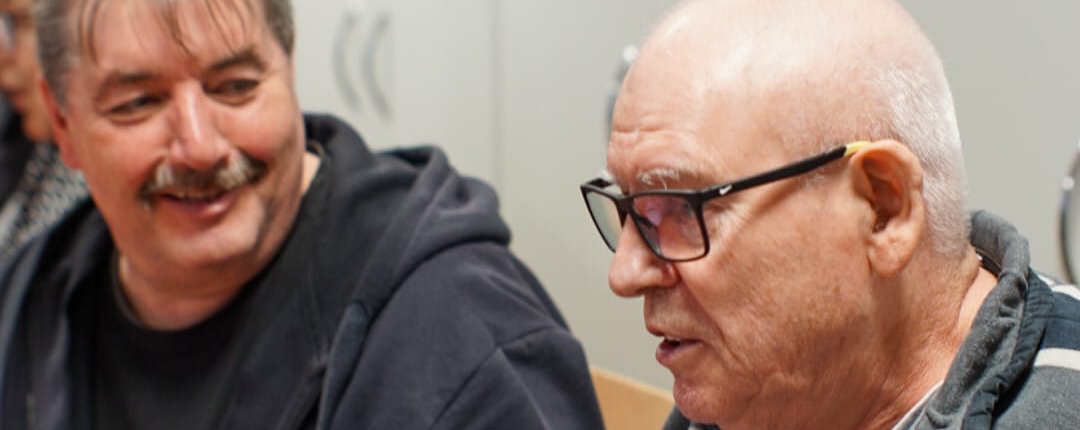 Zwei Männer sitzen an einem Tisch. Einer spricht und gestikuliert. Der andere hört aufmerksam zu.