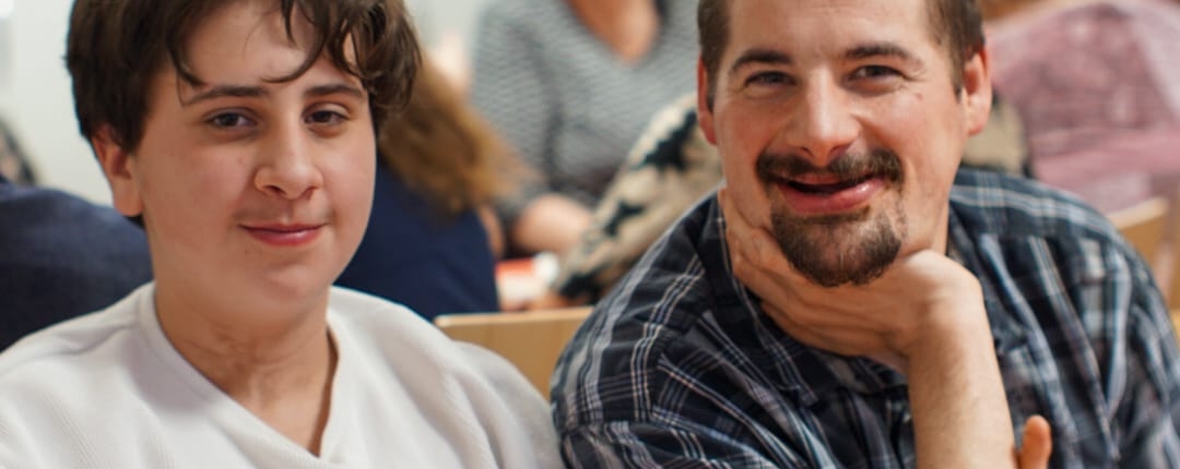 Zwei Personen sitzen an einem Tisch. Ein Mann lächelt, eine Frau schaut in die Kamera. Es stehen Teekannen auf dem Tisch.