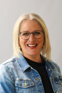 Cueppers_Martina_2026 Porträt einer lächelnden Frau mit blonden, kurzen Haaren und Brille, die ein weißes Oberteil trägt. Der Hintergrund ist neutral und hell.