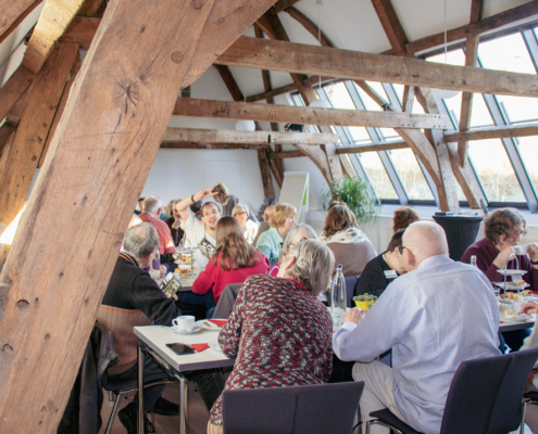 Menschen sitzen an Tischen und essen. Es gibt Essen und Getränke. Die Sonne scheint durch die Fenster.