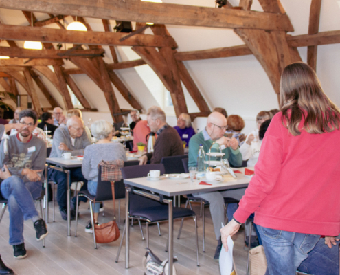 Viele Menschen sitzen an Tischen in einem Raum. Einige stehen auf und unterhalten sich.