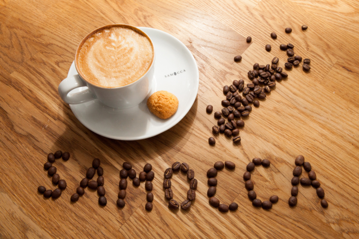 Eine Tasse Kaffee mit einem Keks auf einem Teller. Kaffeebohnen liegen um den Teller.