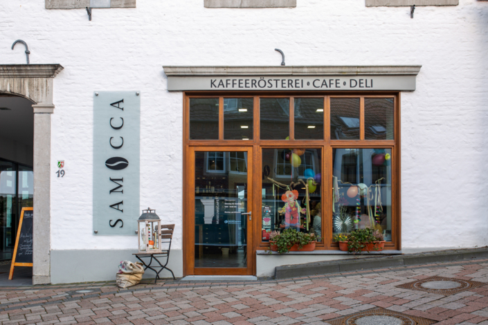 Ein Café mit vielen Pflanzen im Schaufenster. Die Fassade ist weiß und es gibt eine rote Tür.