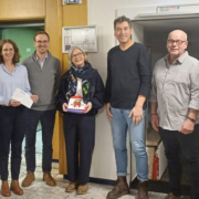 Fünf Personen stehen drinnen. Zwei Frauen und drei Männer. Sie halten Papiere in der Hand.