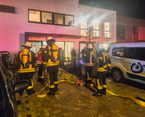 Feuerwehrleute stehen vor einem Haus. Sie tragen Helme und schwere Kleidung. Es ist dunkel draußen.