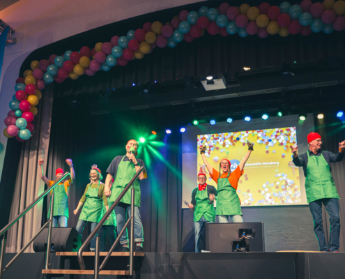 Menschen stehen auf einer Bühne. Sie tragen grüne Kleidung. Im Hintergrund sind bunte Luftballons und ein Bildschirm zu sehen.