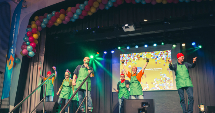 Menschen stehen auf einer Bühne. Sie tragen grüne Kleidung. Im Hintergrund sind bunte Luftballons und ein Bildschirm zu sehen.