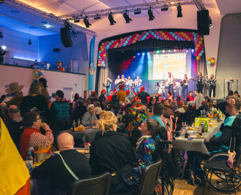 Menschen feiern in einem Saal. Eine Band spielt auf der Bühne. Es gibt Tische mit Essen.