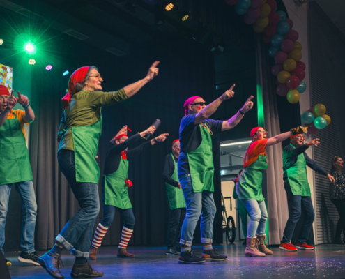 Eine Gruppe tanzende Menschen auf einer Bühne. Sie tragen grüne Westen und bunte Hüte. Es gibt Scheinwerfer und bunte Luftballons.