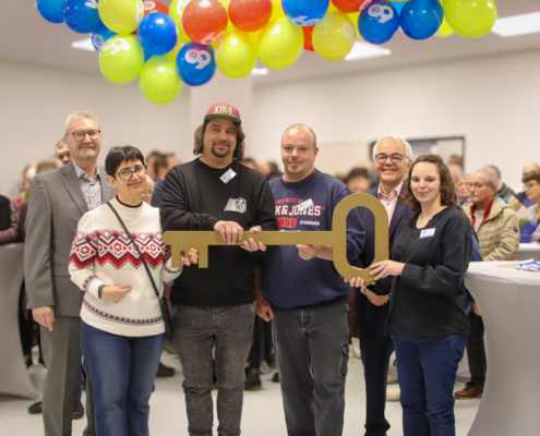 Menschen stehen in einem Raum mit Luftballons. Ein Mann hält einen Schlüssel.