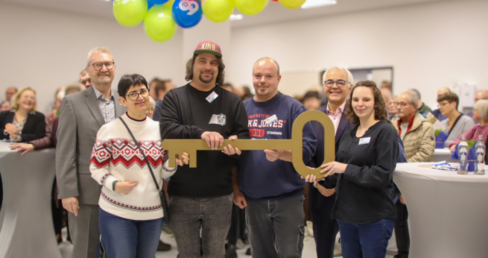 Menschen stehen in einem Raum mit Luftballons. Ein Mann hält einen Schlüssel.