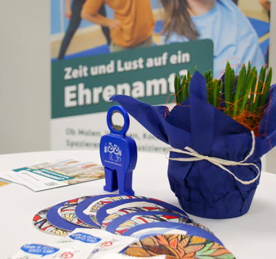 Ein kleiner Topf mit Grünpflanzen steht auf einem Tisch. Daneben liegen blaue Karten und ein blauer Schlüssel.