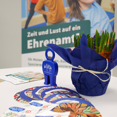 Ein kleiner Topf mit Grünpflanzen steht auf einem Tisch. Daneben liegen blaue Karten und ein blauer Schlüssel.