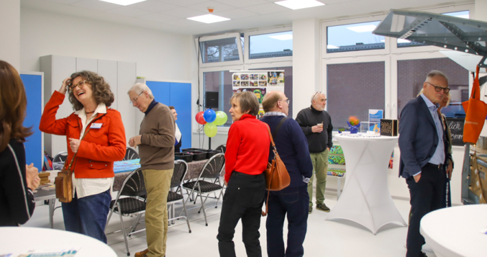 Menschen stehen in einem Raum. Manche sprechen miteinander. Es gibt Tische und Stühle.