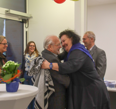 Zwei Personen umarmen sich. Eine Frau mit dunklem Mantel und ein Mann stehen sich gegenüber. Im Hintergrund sind noch andere Leute.