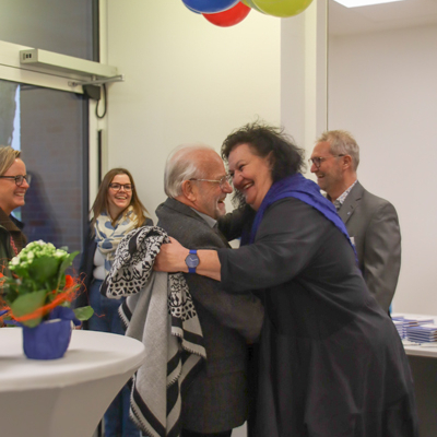 Zwei Personen umarmen sich. Eine Frau mit dunklem Mantel und ein Mann stehen sich gegenüber. Im Hintergrund sind noch andere Leute.