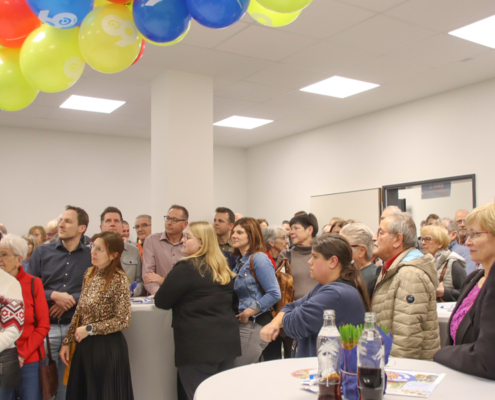 Viele Menschen stehen in einem Raum. Einige sprechen miteinander. Es gibt einen Tisch mit Getränken.