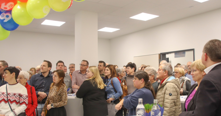 Viele Menschen stehen in einem Raum. Einige sprechen miteinander. Es gibt einen Tisch mit Getränken.