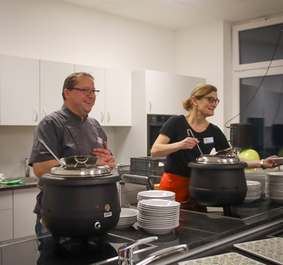 Zwei Personen stehen an einem Buffet. Sie richten Essen an. Es gibt Teller und Töpfe mit Essen.