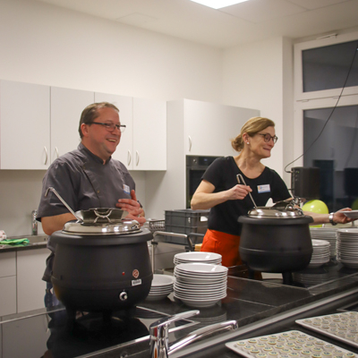 Zwei Personen stehen an einem Buffet. Sie richten Essen an. Es gibt Teller und Töpfe mit Essen.