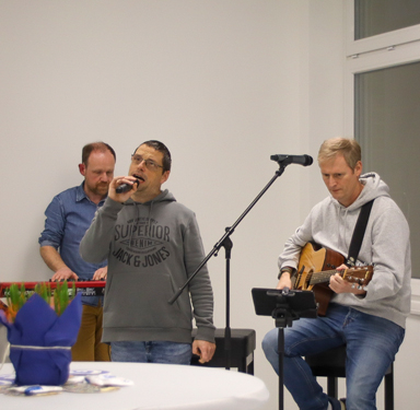 Drei Männer sitzen auf Stühlen. Ein Mann singt in ein Mikrofon. Die anderen beiden spielen Musikinstrumente.