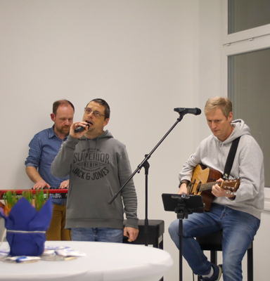 Drei Männer sitzen auf Stühlen. Ein Mann singt in ein Mikrofon. Die anderen beiden spielen Musikinstrumente.